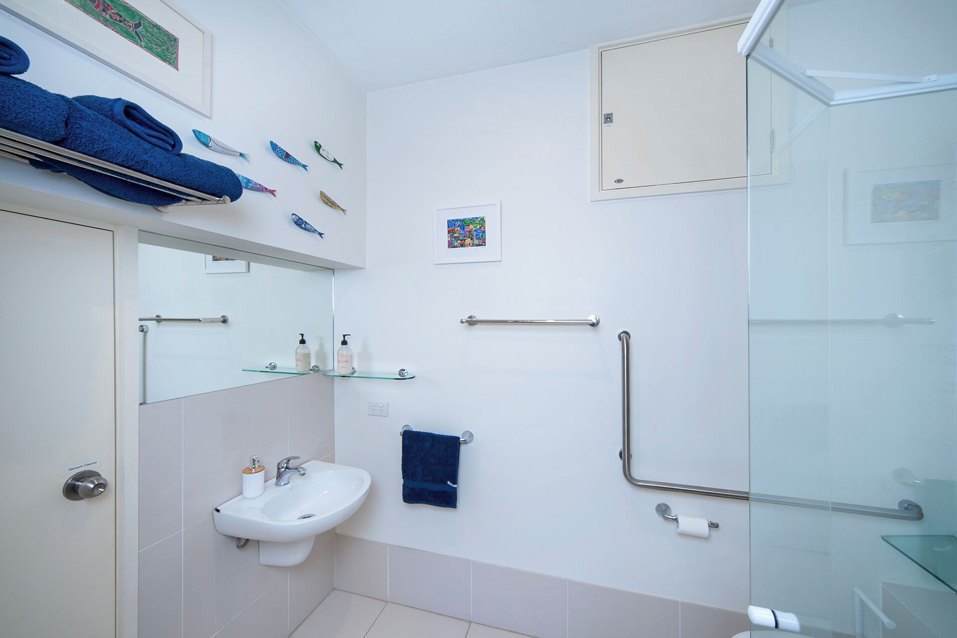 A bathroom with a sink, mirror, towel rack, glass shower enclosure, and handrails.