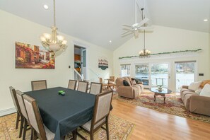 A dining area with a table, chairs, and a chandelier. There is a living room with a sofa and a coffee table. A staircase is visible in the background. - Lake Front Home w/Dock, Kayak, Canoe Right On Lake Hopatcong w/AC Wifi Huge Deck (Jefferson Township)