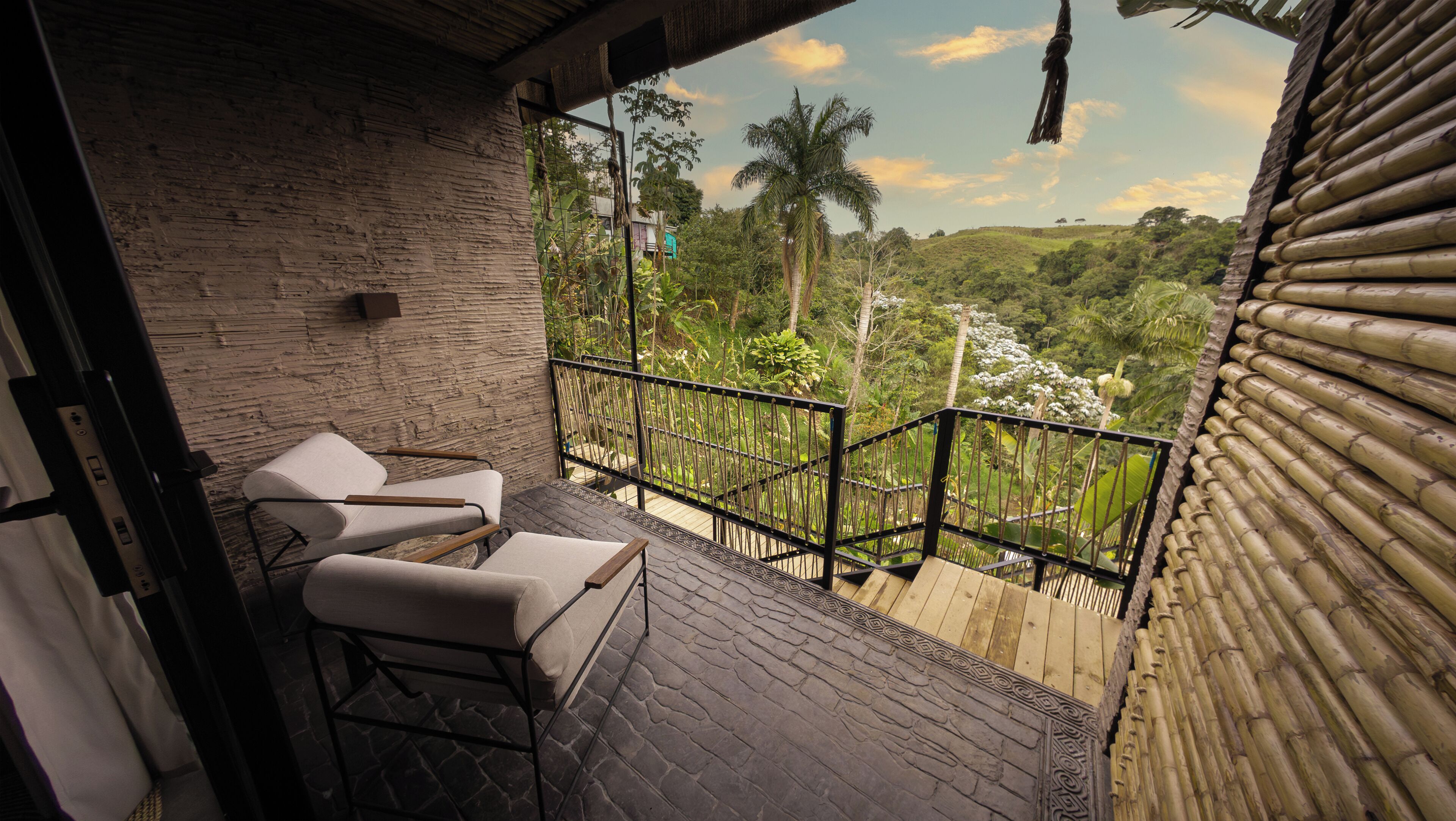 A balcony with two white cushioned chairs and a wooden table, overlooking a lush green landscape with palm trees and a clear sky.