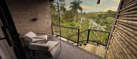 A balcony with two white cushioned chairs and a wooden table, overlooking a lush green landscape with palm trees and a clear sky.