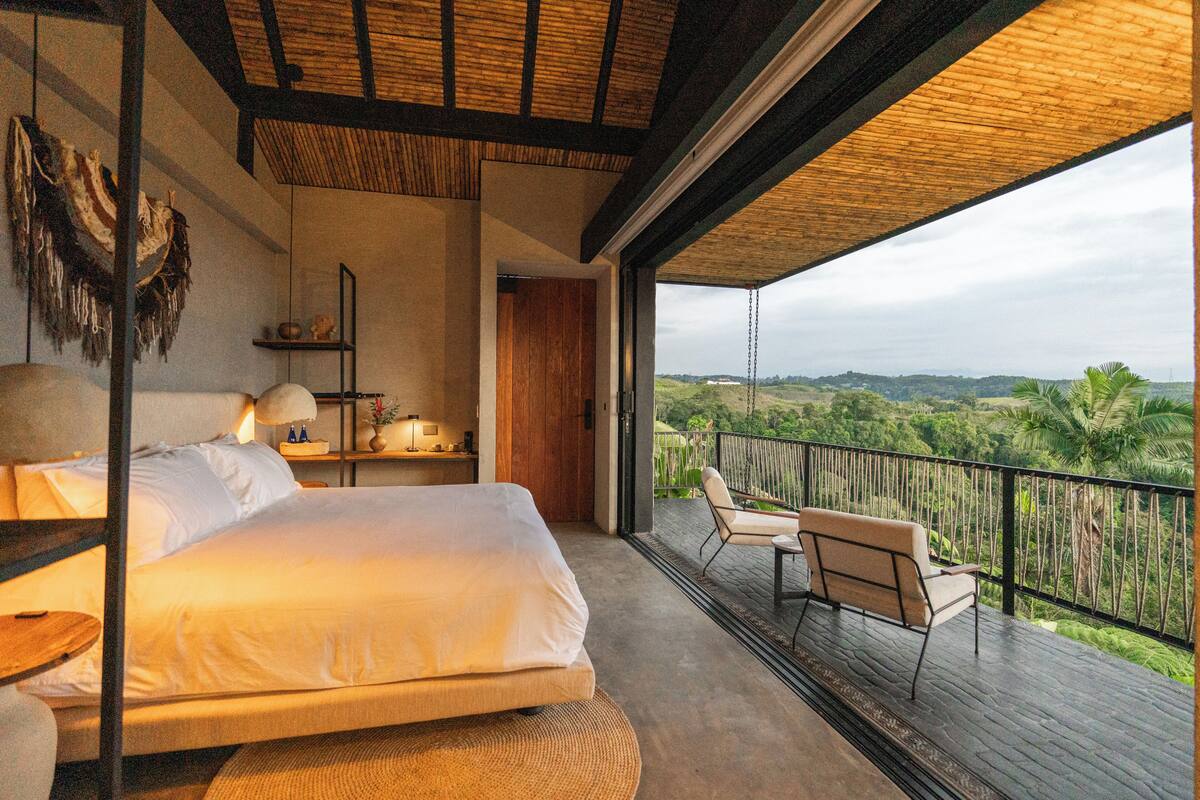 A bedroom with a large bed, bedside tables, and a view of a balcony with chairs.