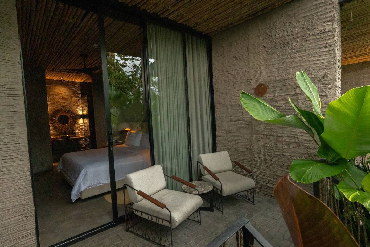 A balcony with a bed, two chairs, and a large plant.