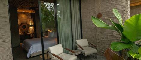 A balcony with a bed, two chairs, and a large plant.