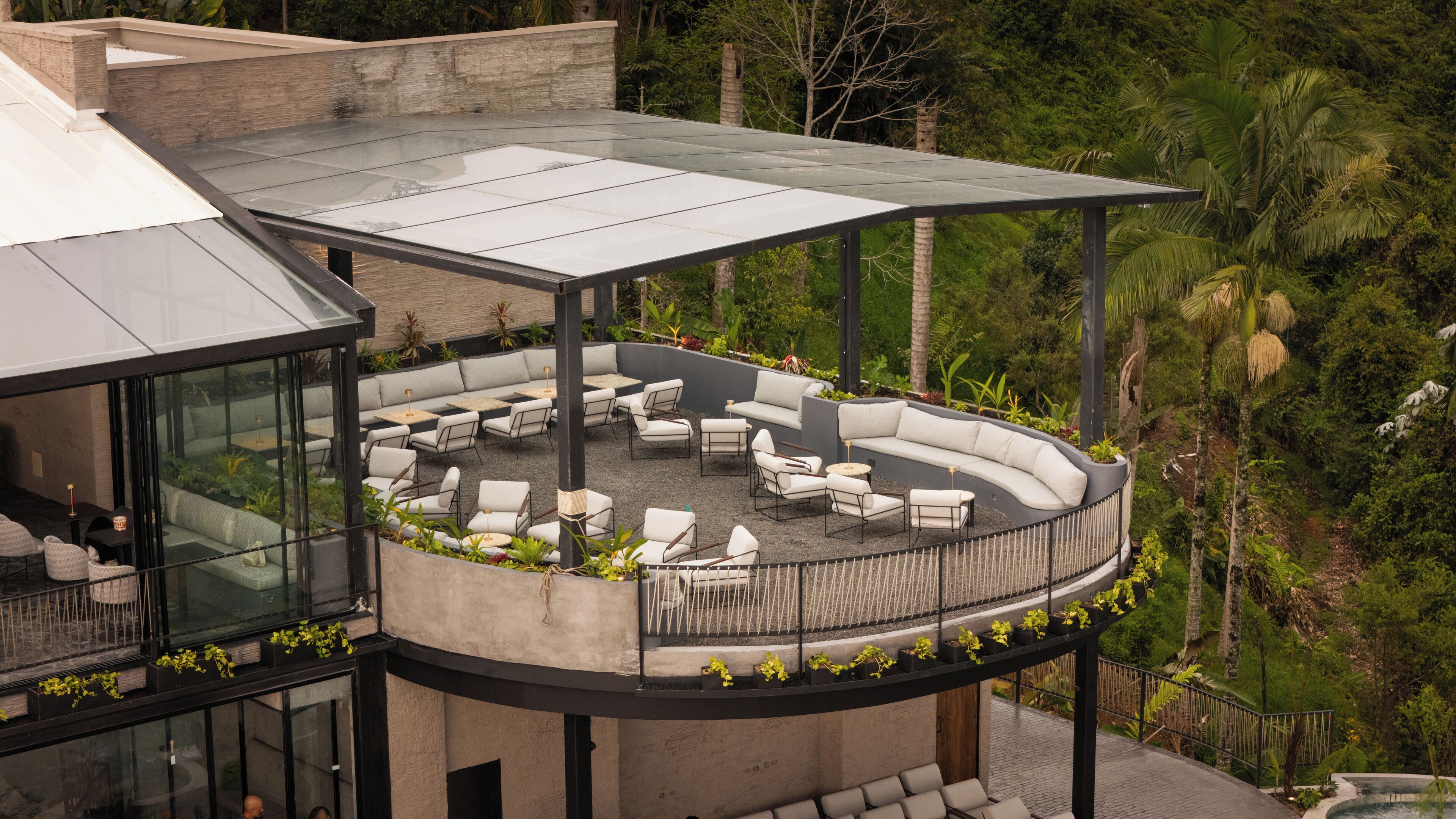 A modern outdoor lounge area with a glass roof, cushioned seating, and a surrounding garden.