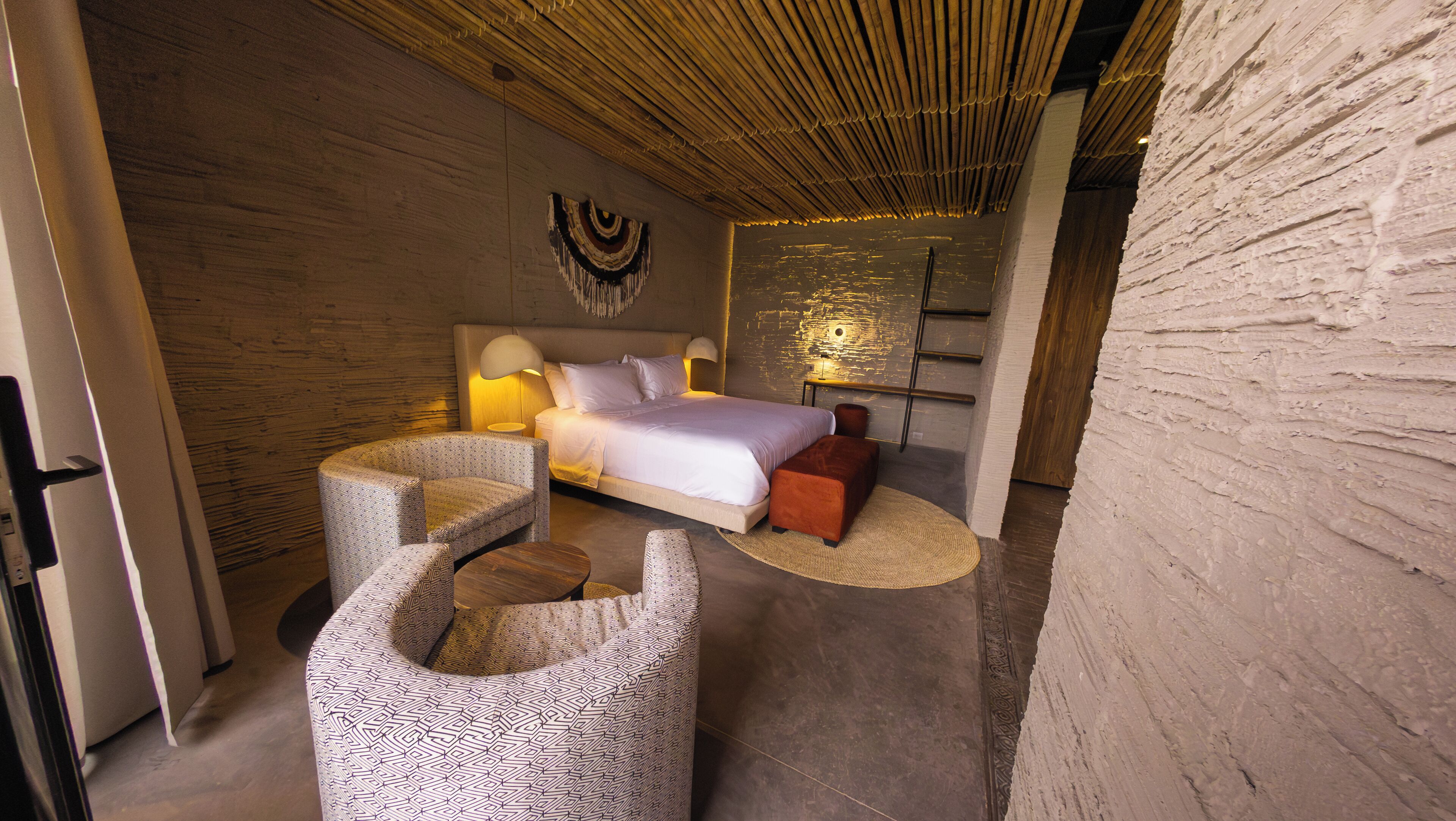 A modern bedroom with a large bed, two chairs, a small table, and a wall-mounted lamp.