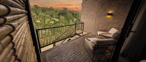 Un balcon avec vue sur une végétation luxuriante et un coucher de soleil. On y trouve deux chaises blanches et une petite table.