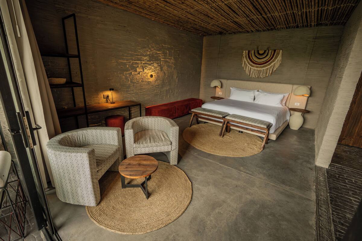 A modern hotel room with a large bed, two armchairs, a small round table, and a wooden bench.