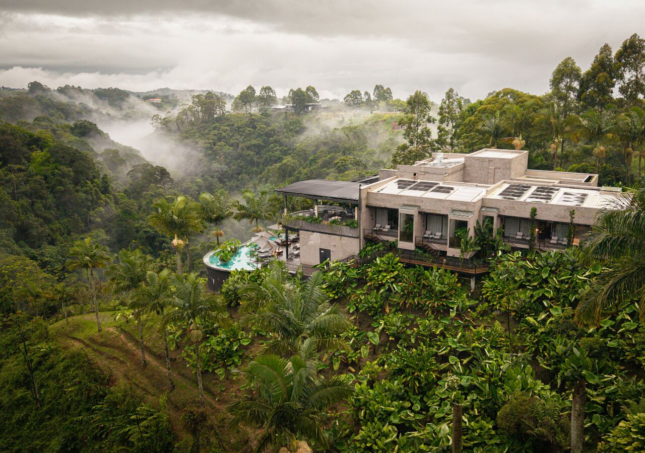 A modern hotel with a pool, surrounded by lush greenery and a mountainous backdrop.