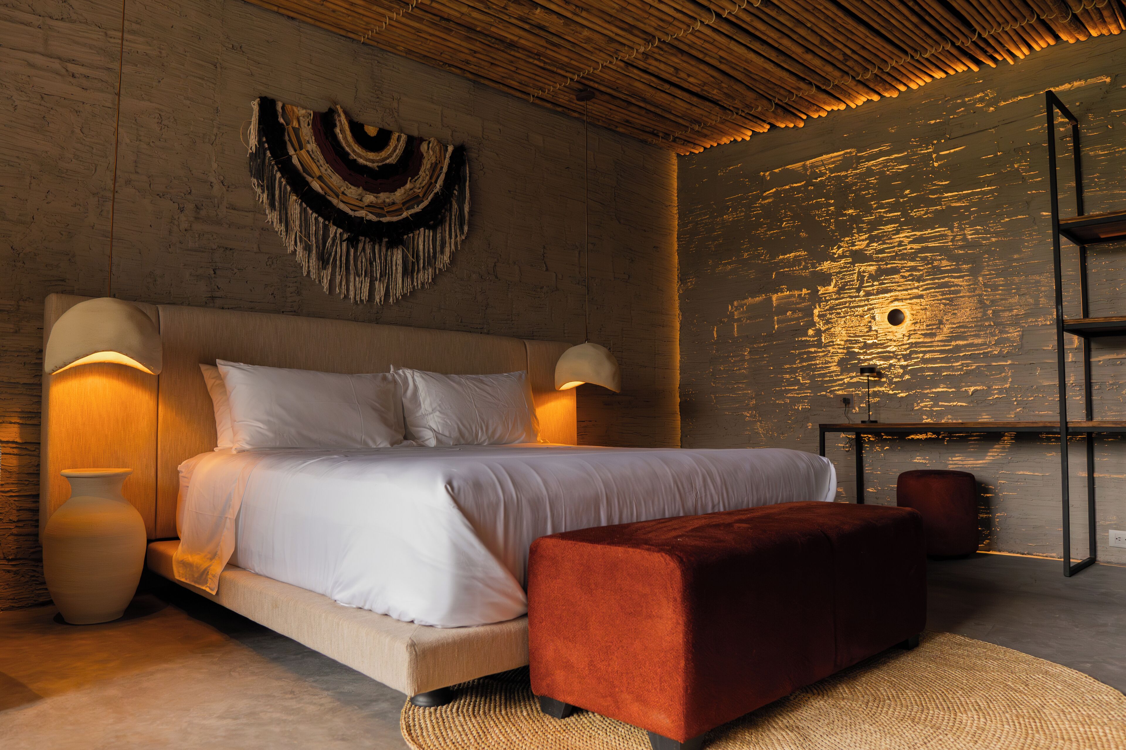 A modern bedroom with a bed, two bedside lamps, a red ottoman, a woven wall hanging, and a wooden shelf.