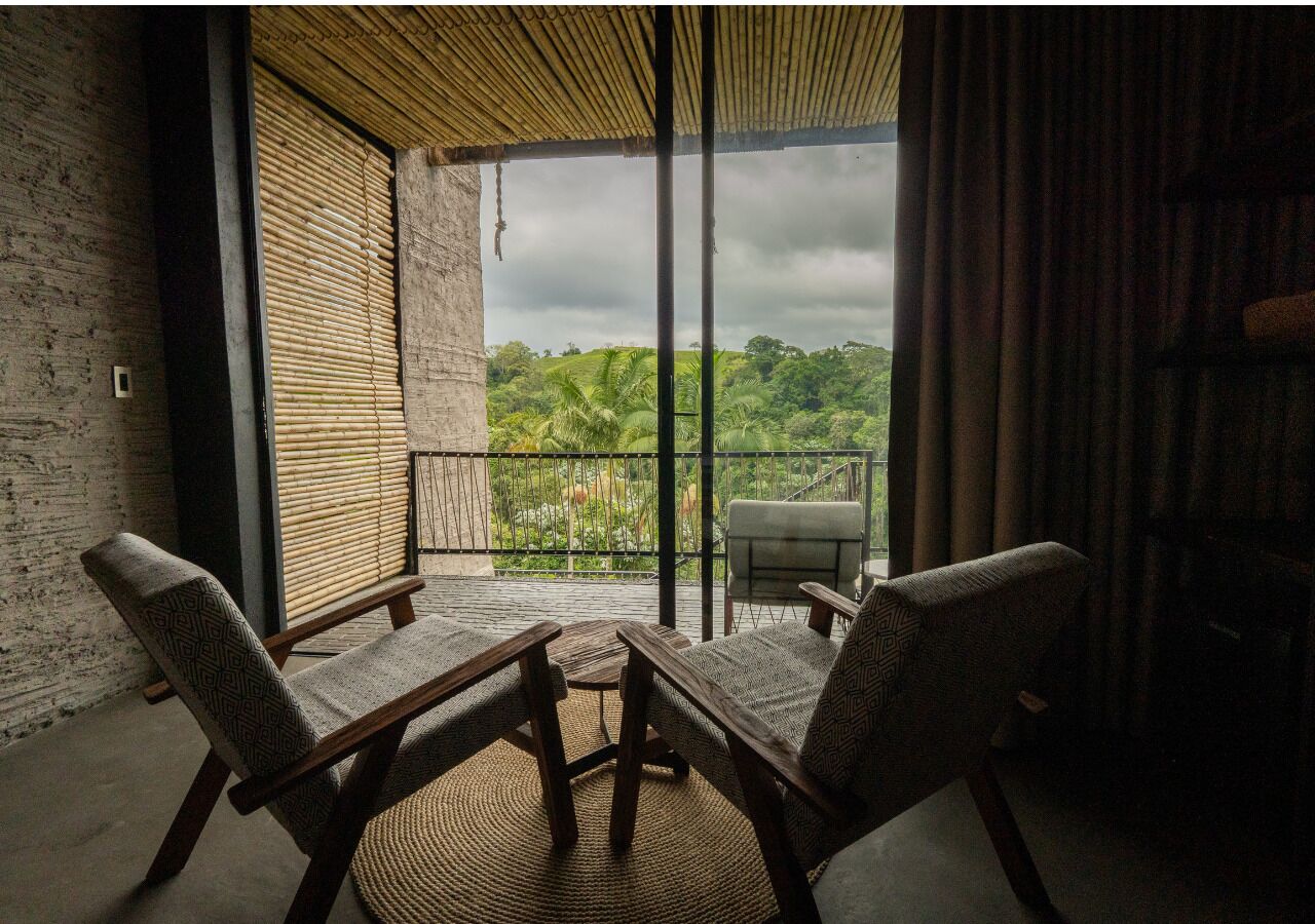 A balcony with two chairs and a table, a view of lush greenery, and a bamboo ceiling.