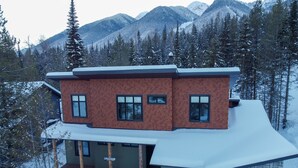 Une maison moderne à deux étages, dont le toit est recouvert de neige, entourée d’une forêt et de montagnes.