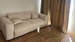 Un canapé beige, une table basse en verre, dans une pièce au plancher en bois et dotée d’une fenêtre avec des rideaux.