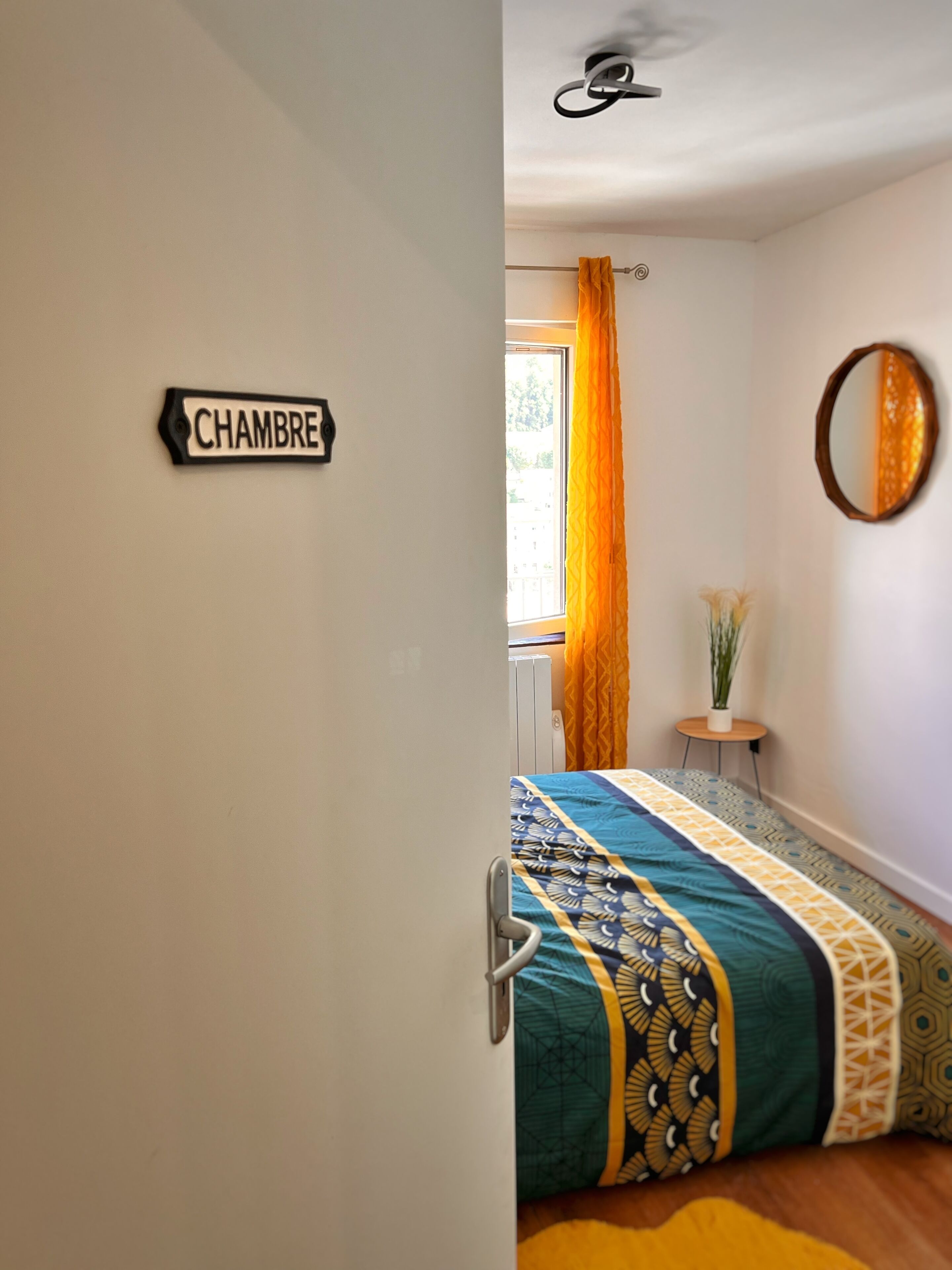 Un dormitorio con cama, ventana con cortinas, una mesita con planta y un espejo en la pared.
