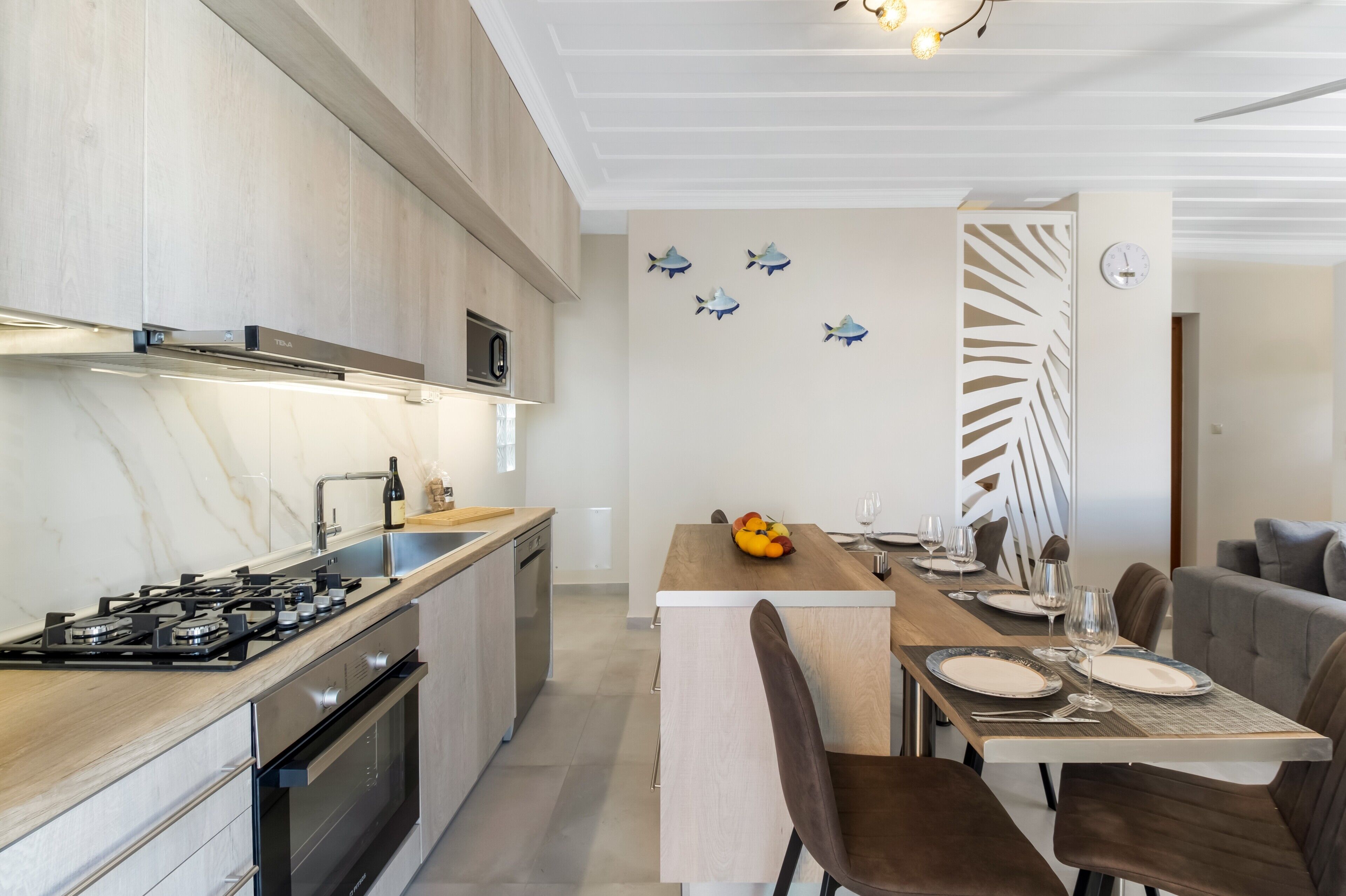 A modern kitchen with a gas stove, microwave, and wooden cabinets. There is a dining area with a table set for two, a decorative wall with fish decals, and a clock.