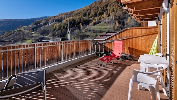A wooden deck with a view of a mountain and a church, white plastic chairs, and a red chair. - Apartment "Haus Peer A 2" with Mountain View, Private Terrace, and Wi-Fi (Mals)