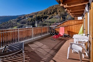 Eine Holzterrasse mit Blick auf einen Berg und eine Kirche, weiße Plastikstühle und ein roter Stuhl.