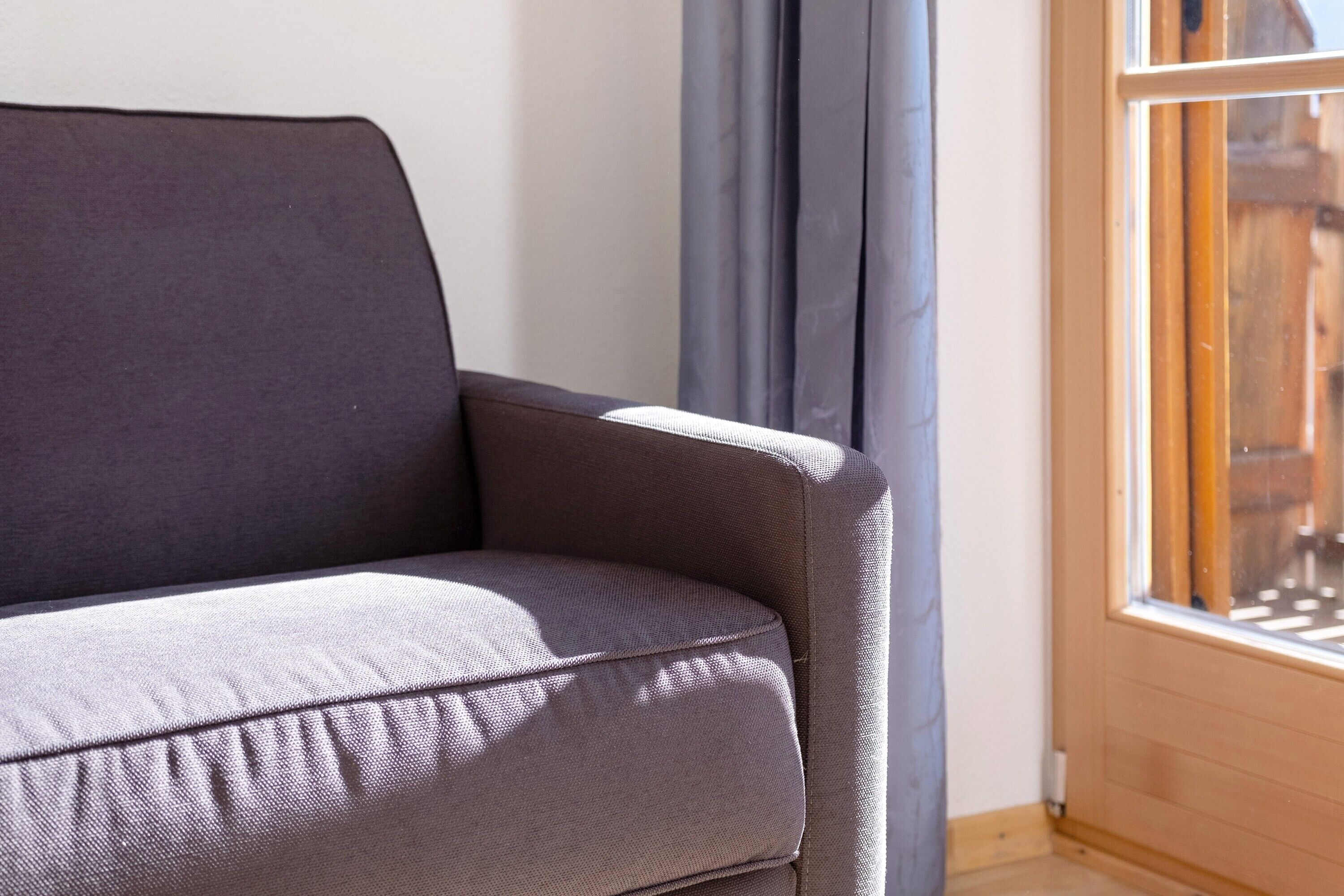 A corner of a room with a grey sofa, a window with wooden frames, and a curtain.
