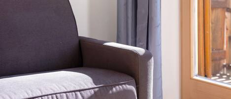 A corner of a room with a grey sofa, a window with wooden frames, and a curtain.