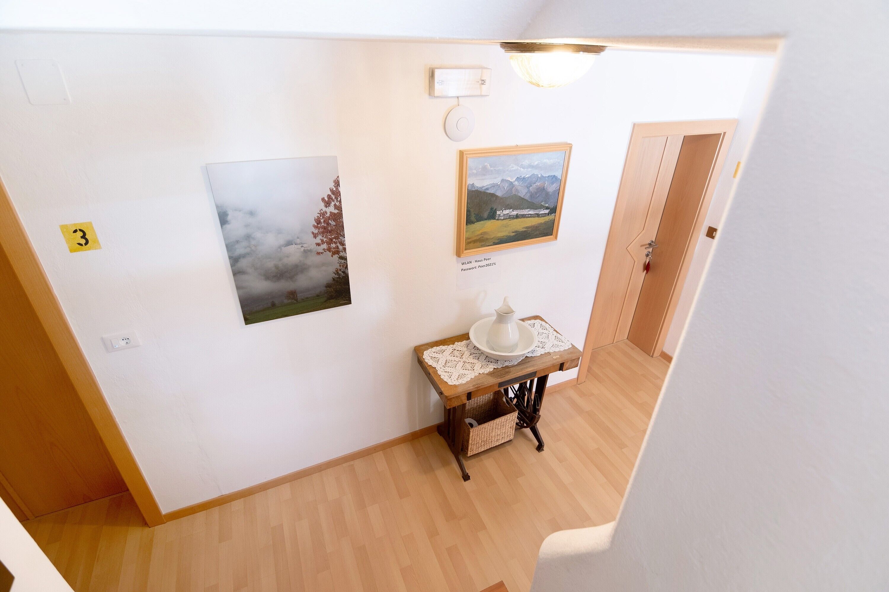 A hallway with a wooden floor, a table with a white vase, and two framed pictures on the wall.