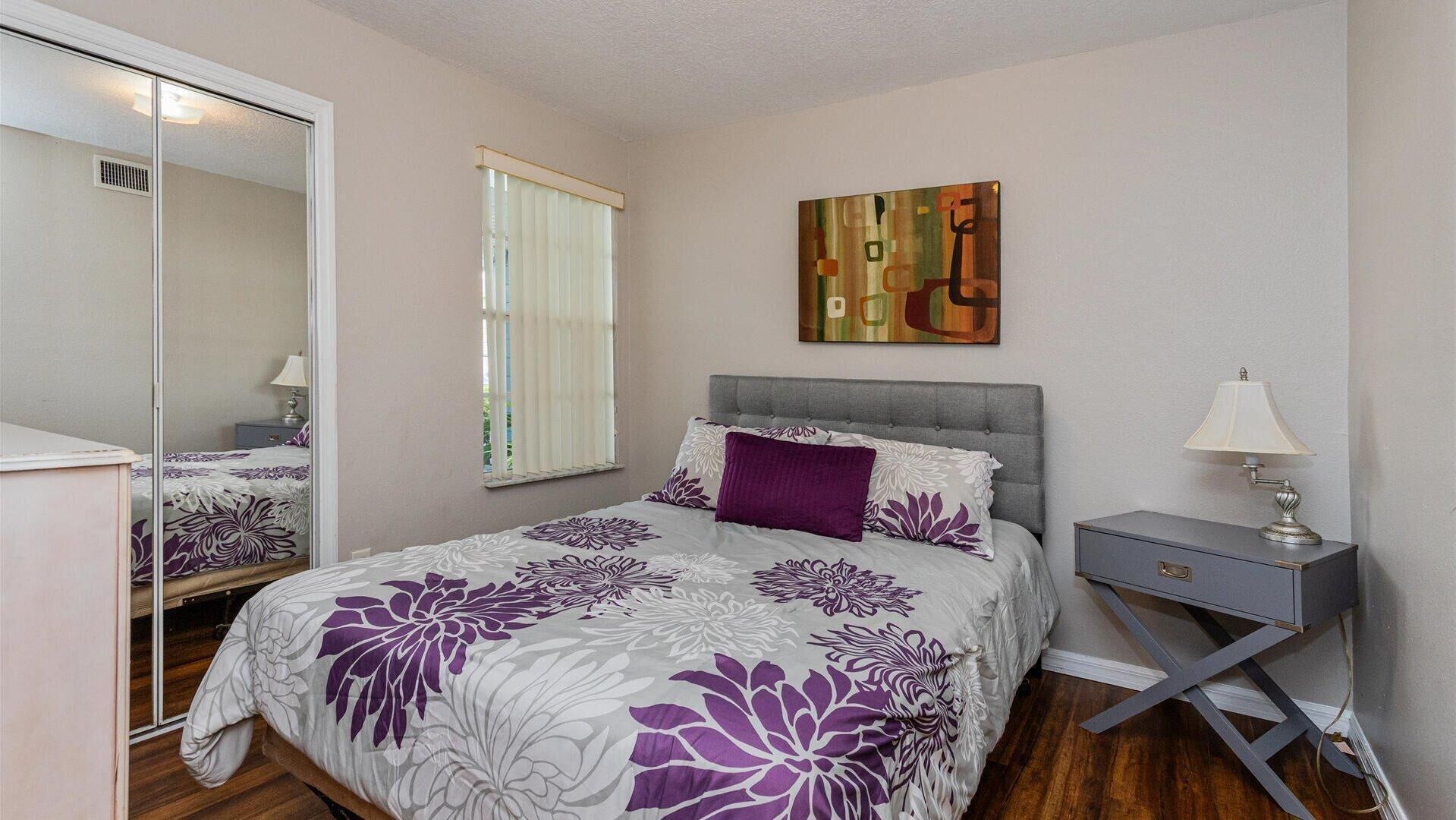 A bedroom with a bed, a bedside table, a lamp, a mirror, and a window with blinds.