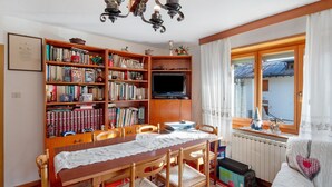 A cozy living room with a wooden dining table, bookshelves, a television, and a window with curtains. - Apartment 'Mulaz' with Mountain View, Shared Garden and Wi-Fi (Falcade)
