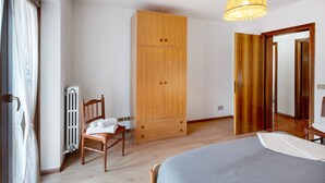 Een slaapkamer met een bed, een houten garderobe, een stoel, een radiator en een deur naar een andere kamer.
