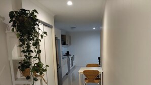 A modern, minimalist hallway with a dining area featuring a white table and wicker chairs. A large potted plant is visible on the left, and a wicker bench is on the right. The kitchen area is visible in the background with white cabinets and a stove.