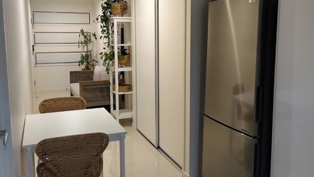 A modern kitchen with white cabinets, a stainless steel refrigerator, a dining table with wicker chairs, and a built-in shelving unit with baskets.