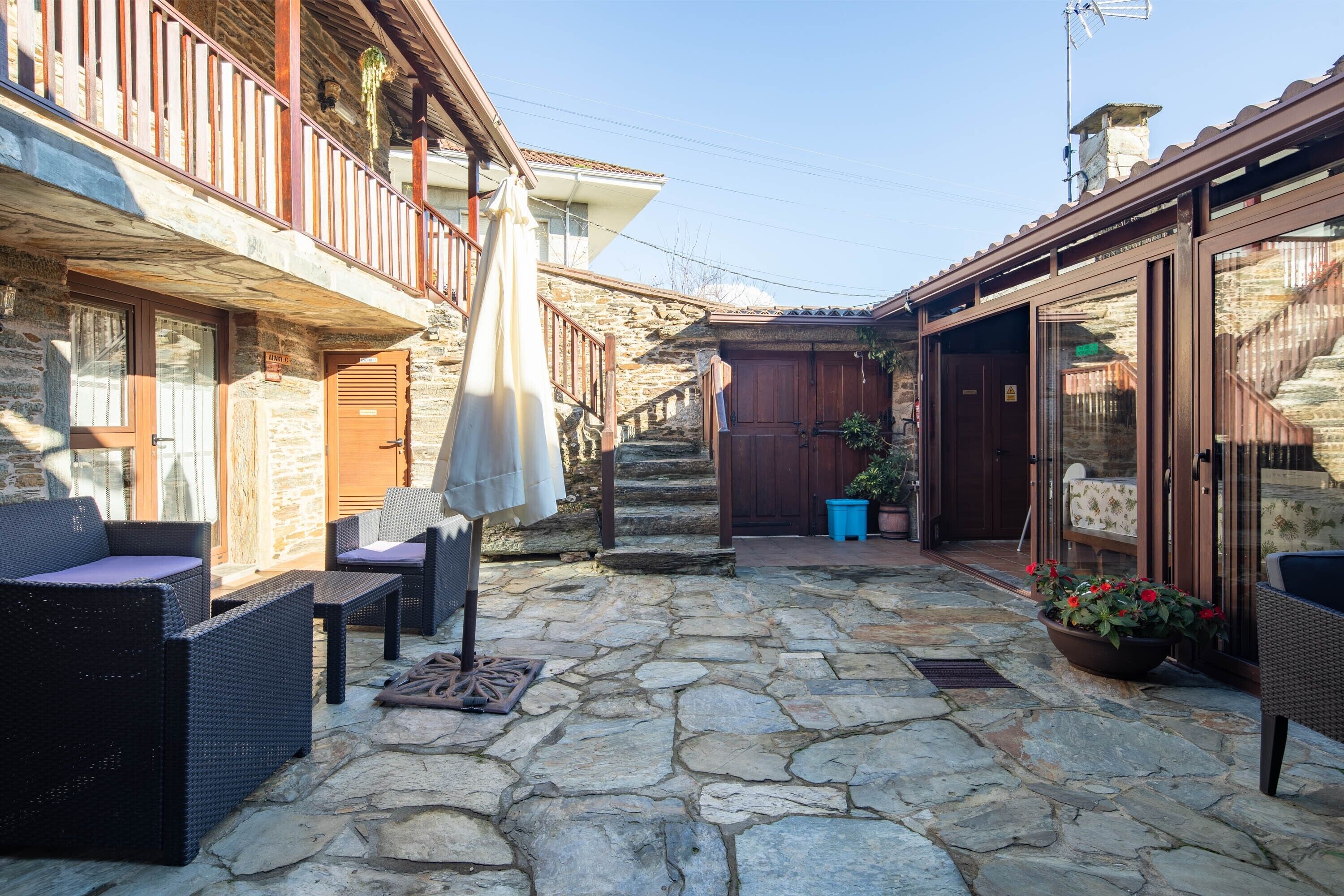 A stone-floored courtyard with wicker furniture, a wooden staircase, and a view of the surrounding buildings.