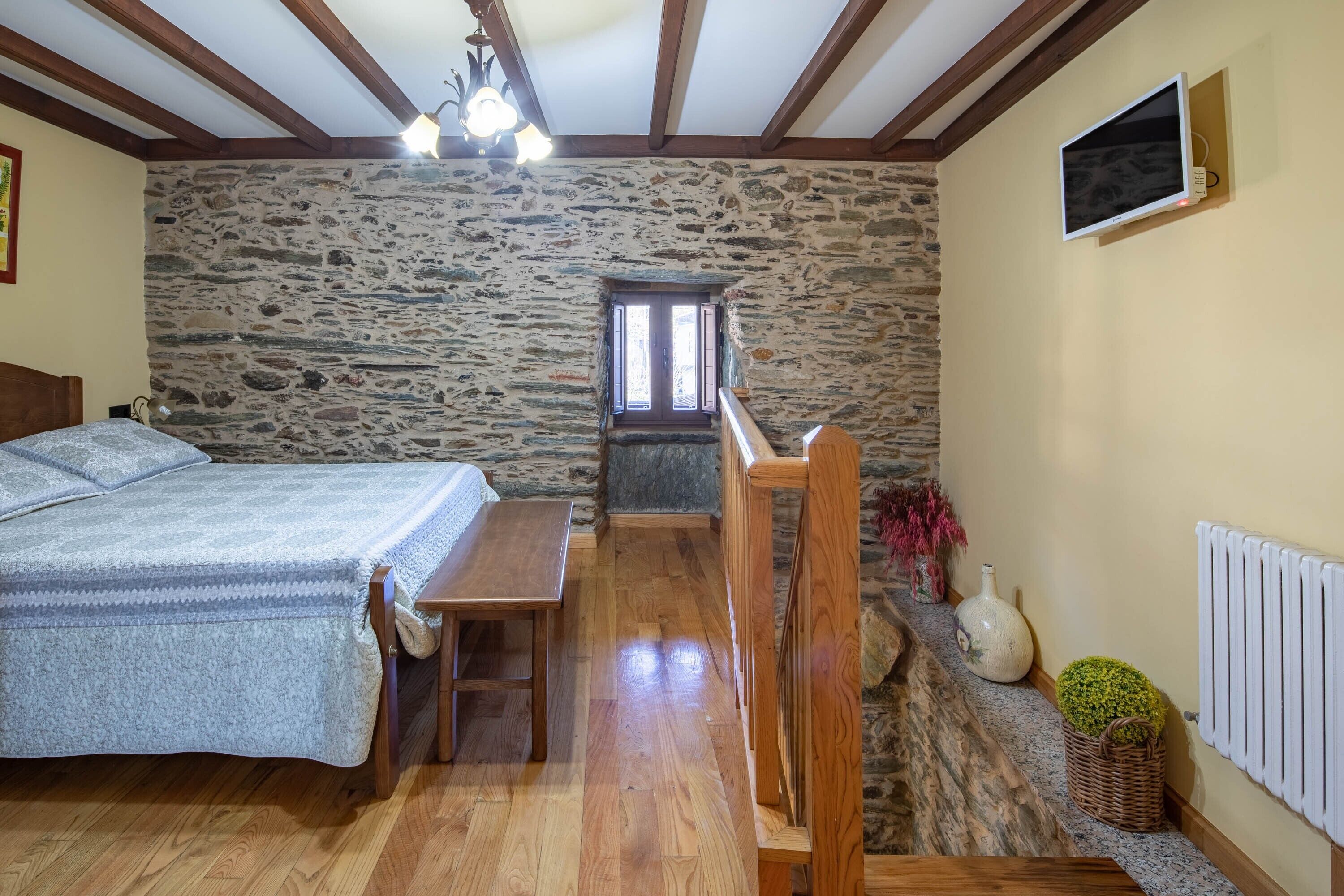 A bedroom with a stone wall, wooden ceiling beams, a bed, a wooden bench, a small table, a television, and a radiator.