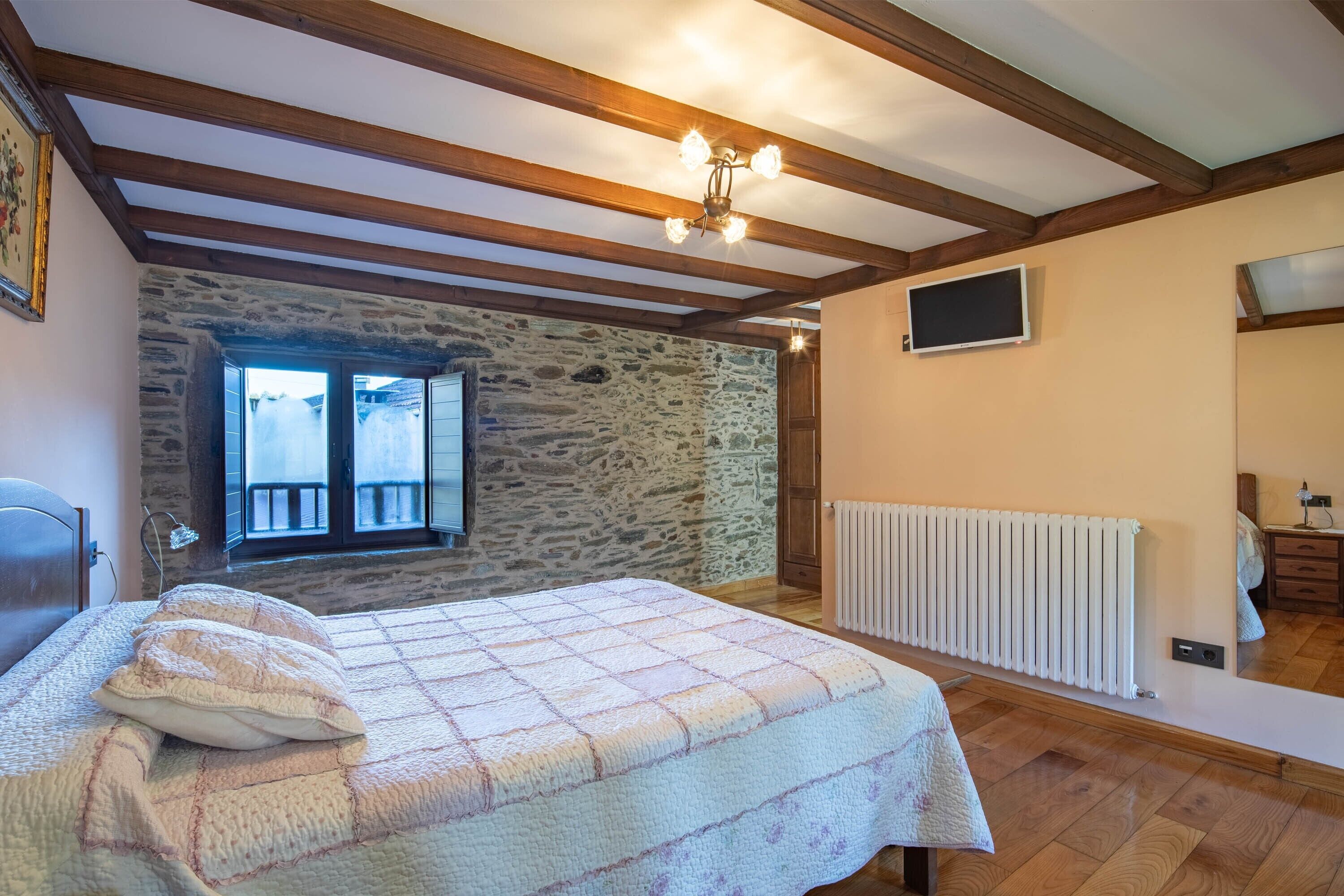 Une chambre avec un mur en pierre, des poutres en bois, un lit recouvert d’une courtepointe, un radiateur et un téléviseur fixé au mur.