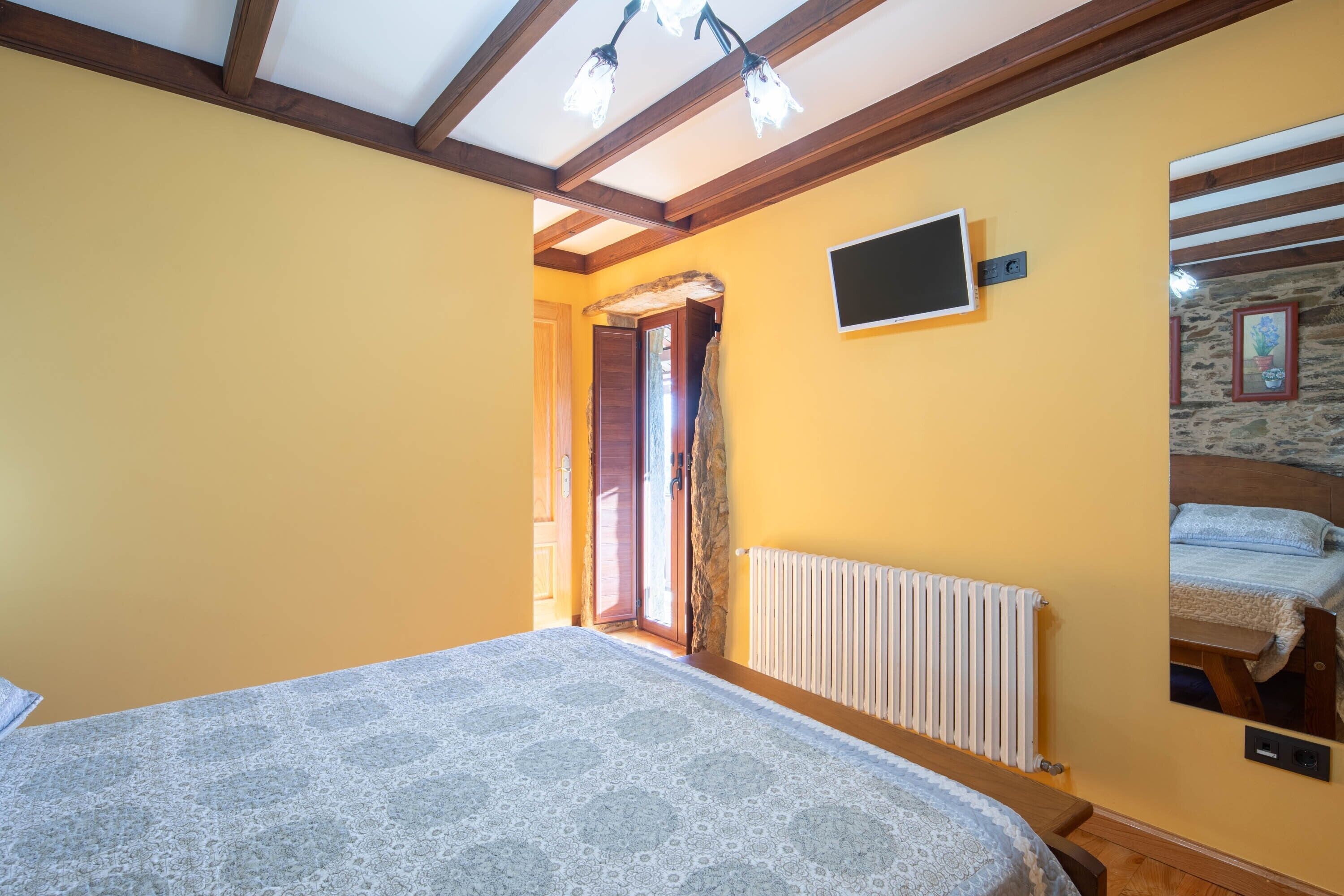 A bedroom with a bed, wooden ceiling beams, a radiator, a television, and a stone wall.