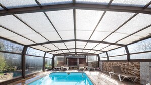 A covered swimming pool with a glass ceiling, lounge chairs, and a stone wall.