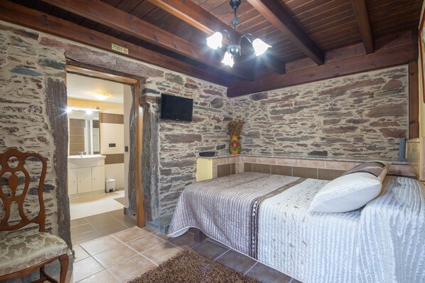 A bedroom with a stone wall, wooden ceiling beams, a bed with a white bedspread, a wooden chair, and a television on the wall.