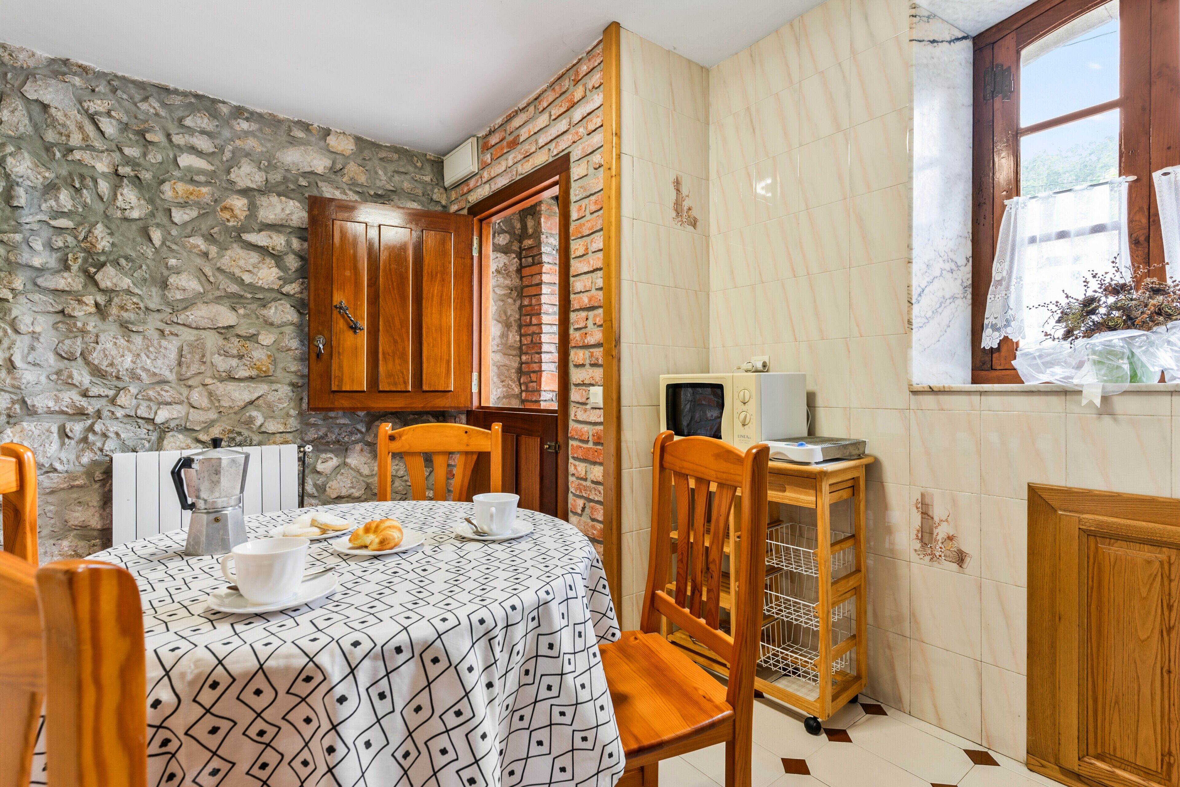 Uma área de refeições com parede de pedra, armários de madeira, parede revestida de azulejos, uma janela com cortinas de renda, uma cafeteira, uma mesa com toalha estampada, cadeiras e um carrinho pequeno com um computador.