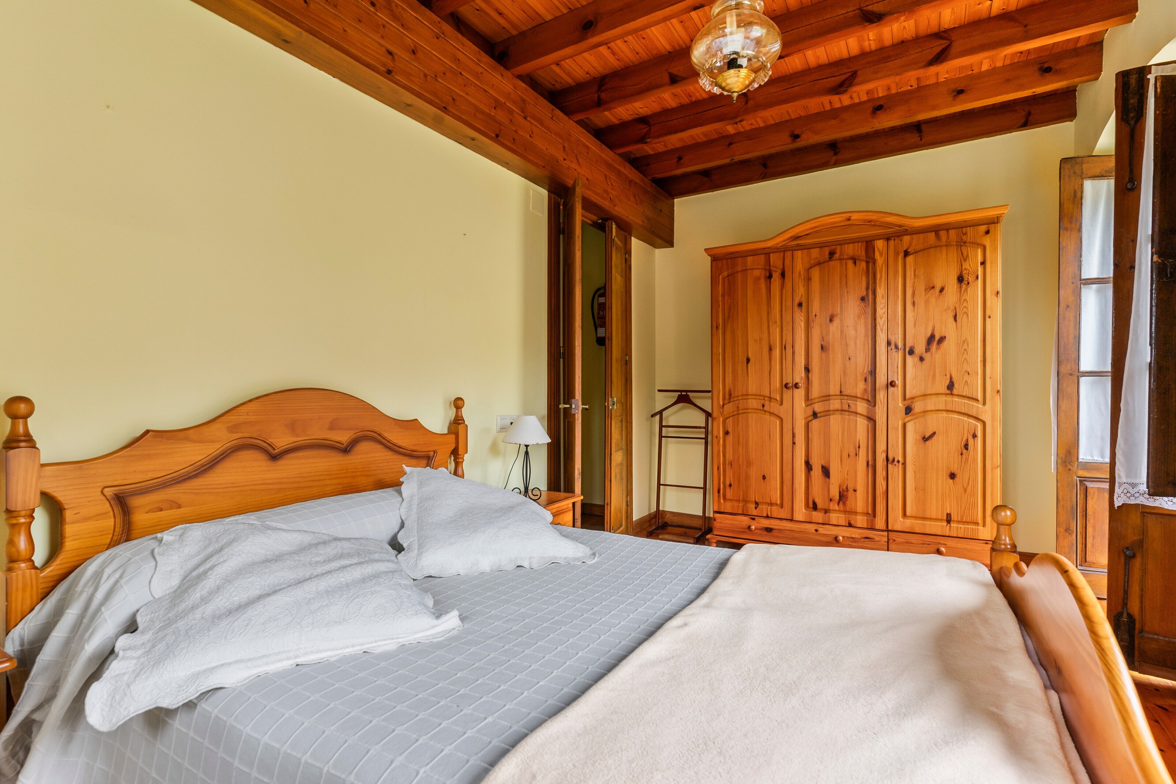 A wooden bed with a headboard, a nightstand, and a wardrobe in a room with wooden beams on the ceiling.