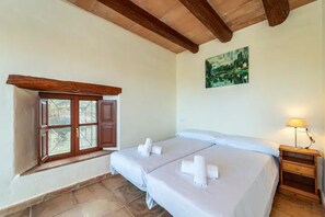 A bedroom with two beds, a window with wooden shutters, a painting on the wall, and a bedside table with a lamp.