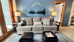 A living room with a grey sofa, patterned cushions, two black ottomans, and a wall mural of mountains and trees.