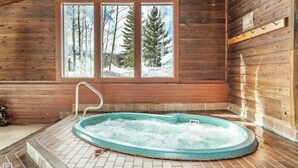 A wooden cabin with a hot tub in the center, large windows with a snowy landscape view, and a tiled floor.