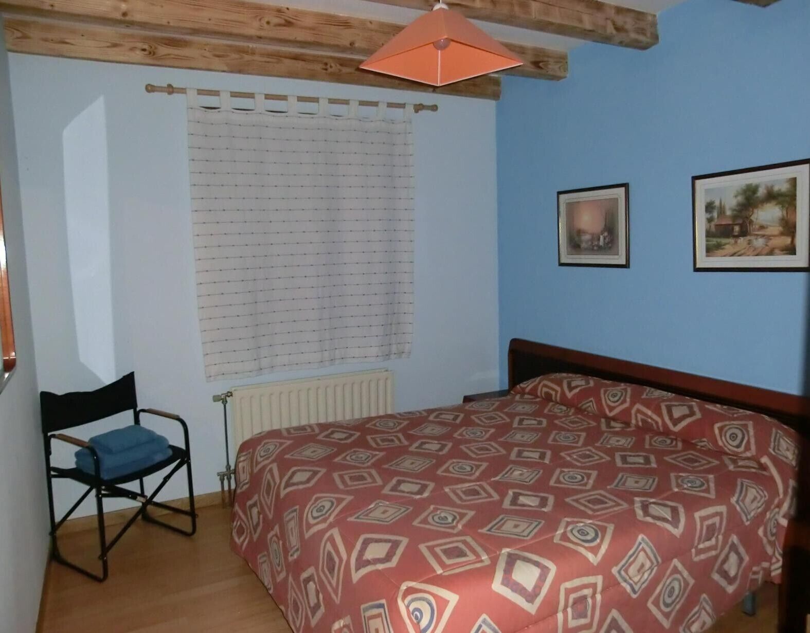 Un dormitorio con cama, una silla, un radiador, una ventana con persianas y dos cuadros en la pared.