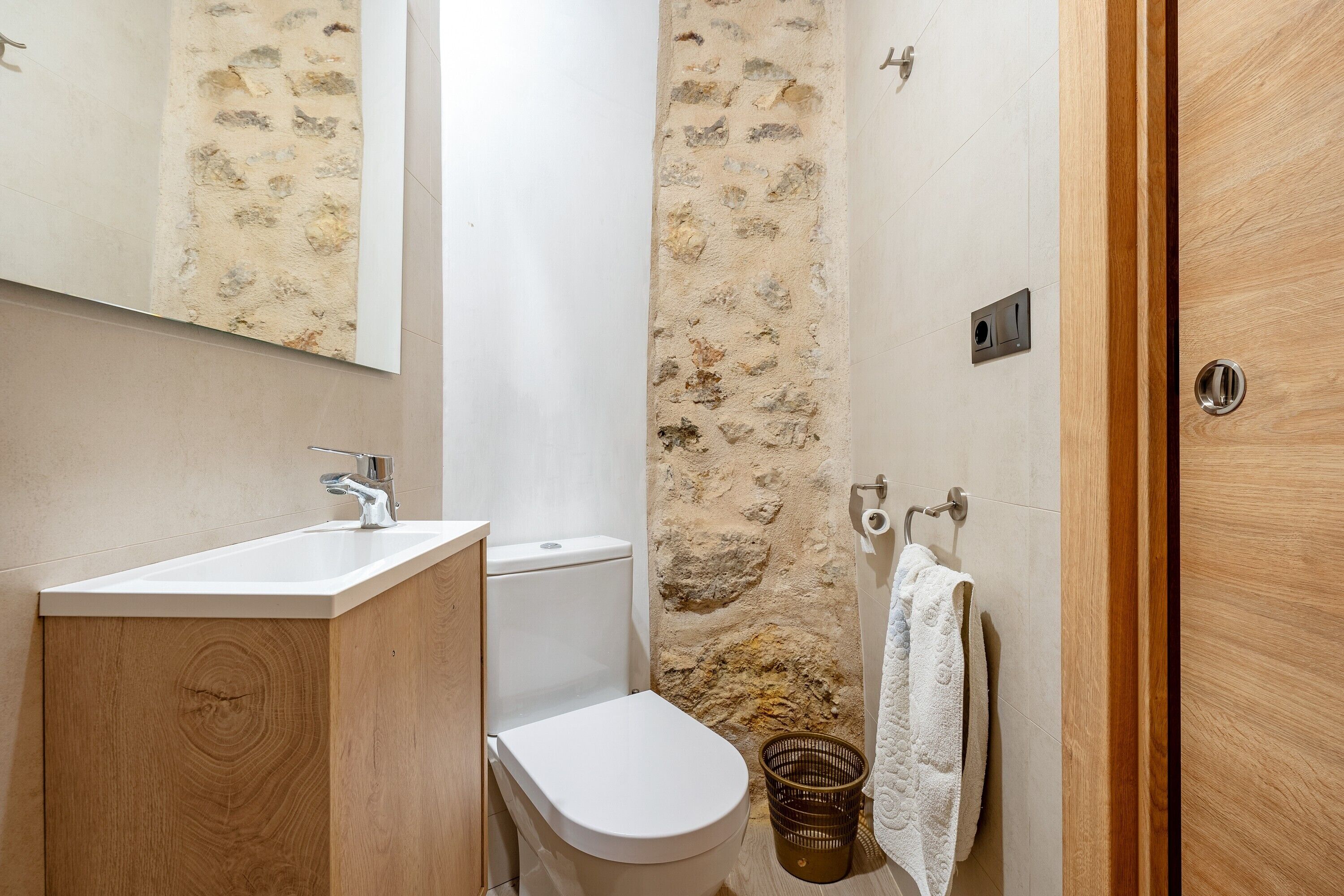 A bathroom with a wooden vanity, white sink, toilet, and a stone shower wall.