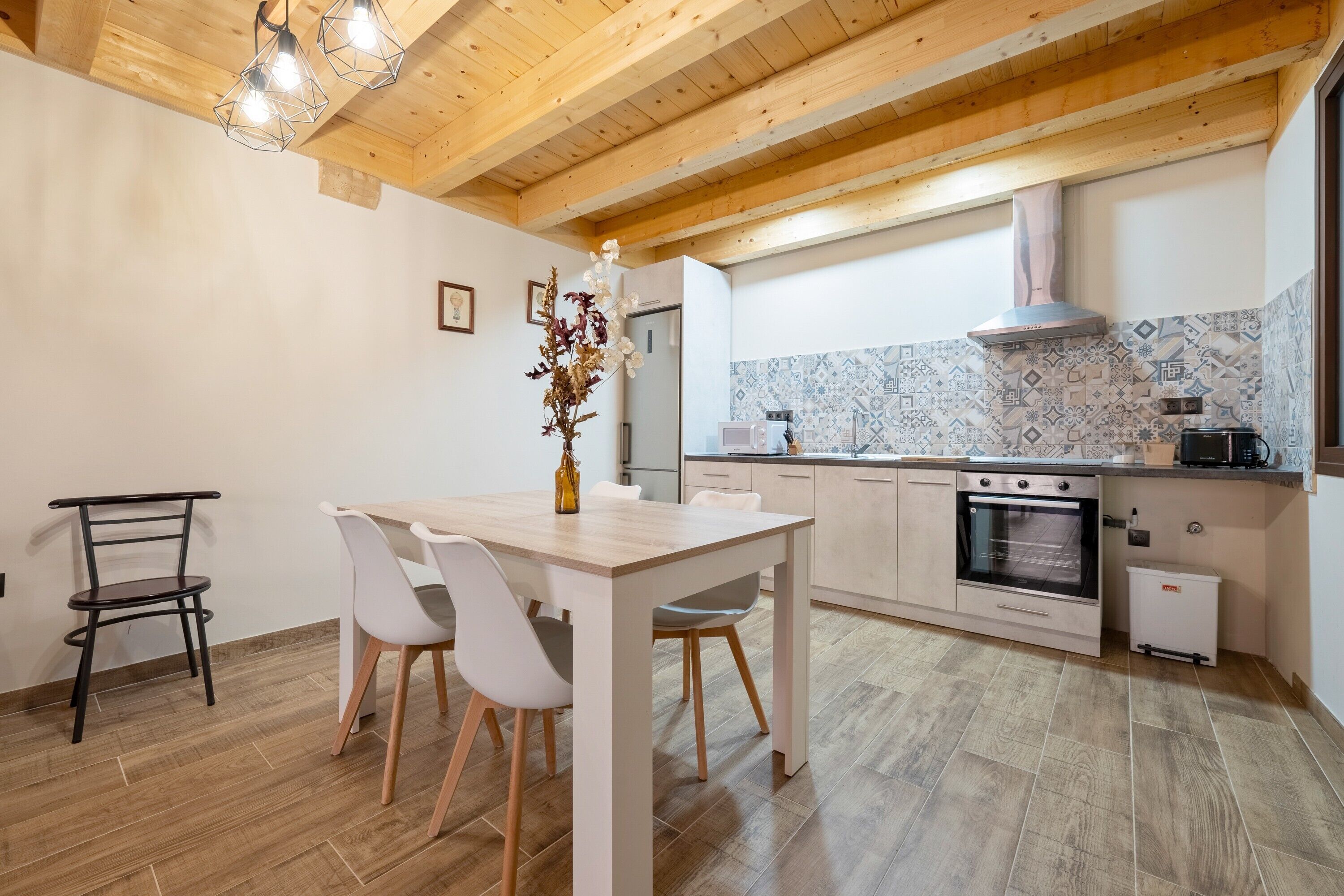 A modern kitchen with wooden beams, a dining table with chairs, a built-in oven, and a patterned backsplash.