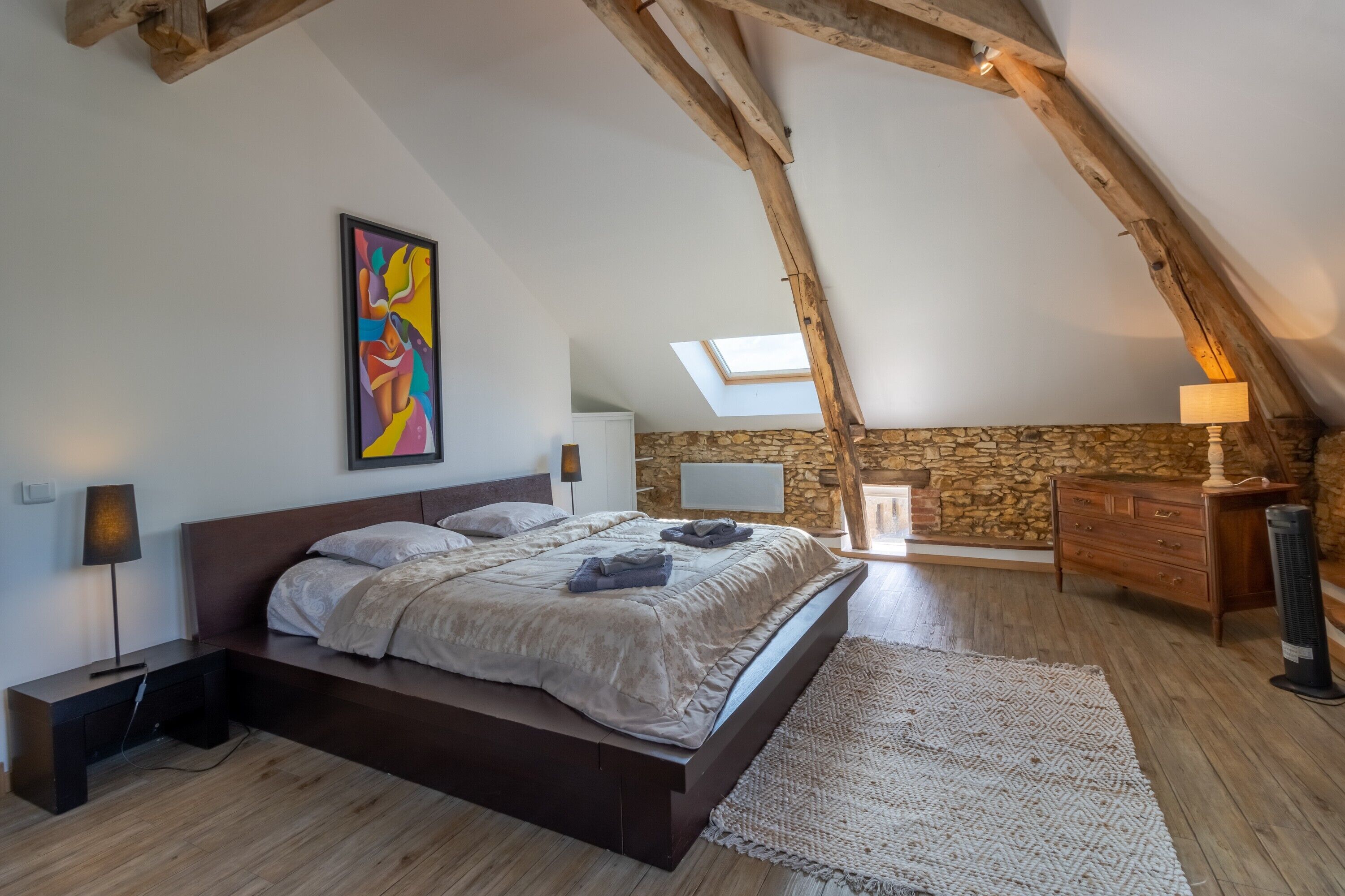 A bedroom with a sloped ceiling, a bed, a stone wall, a skylight, a dresser, and a lamp.