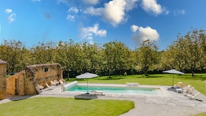 Zona de piscina con sillas de descanso y sombrillas, rodeada por un muro de piedra y árboles.