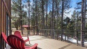 A wooden deck with red Adirondack chairs, overlooking a forested area with a view of a cabin. - 'Enchanted Pines' Mountain Escape! (Angel Fire)