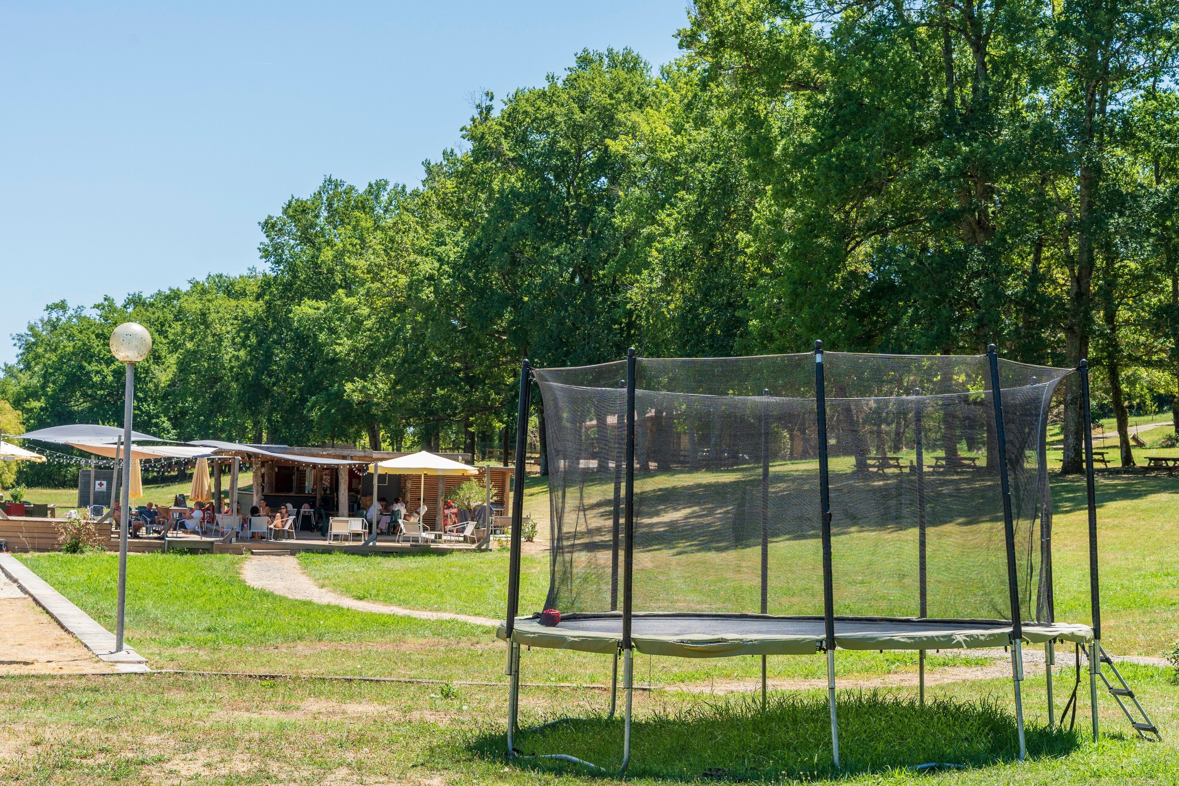 A park with a trampoline enclosed by a safety net, surrounded by trees and a picnic area with tables and chairs.