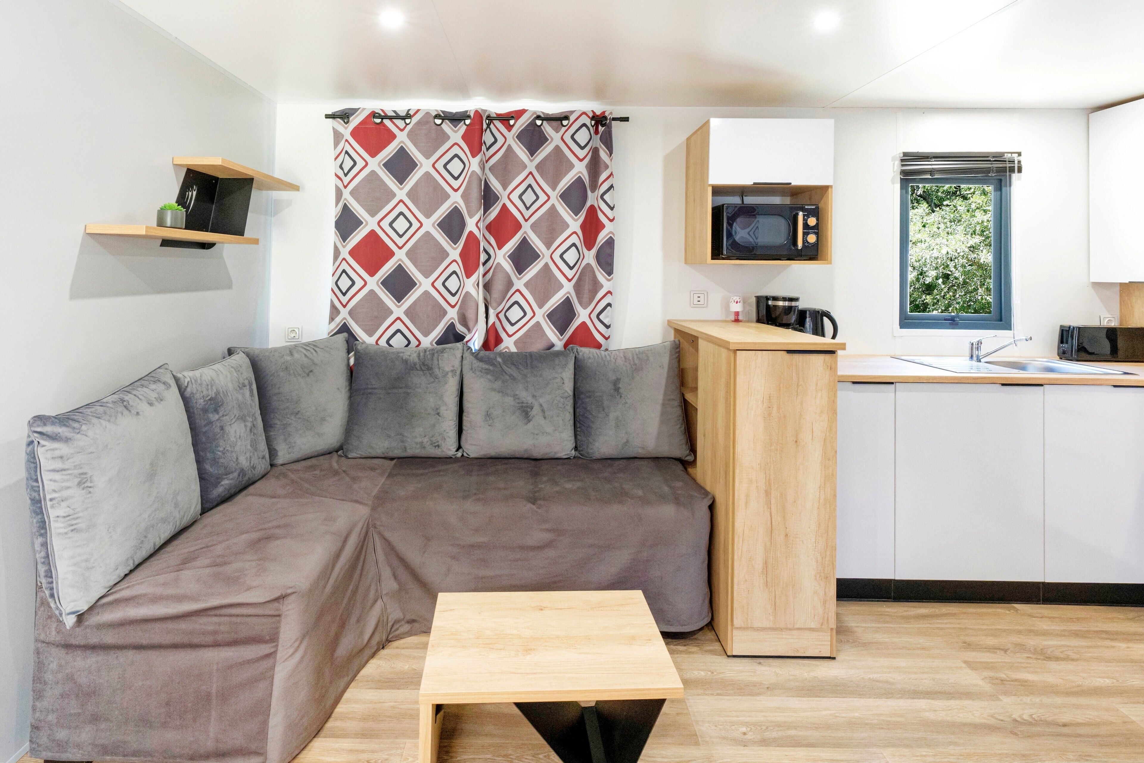 Una kitchenette moderna con microondas, pileta y ventana. Un sofá seccional marrón con cojines grises está ubicado frente a una cortina estampada. Una pequeña mesa de madera se encuentra frente al sofá.