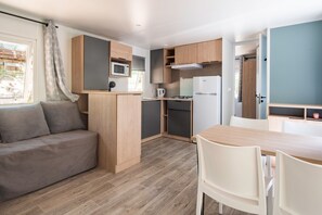 A compact kitchenette with a microwave, stove, and refrigerator. There is a wooden dining table with chairs and a sofa with cushions. - Mobile Home 'Mobilhome Confort 4 Personnes 64' with Private Terrace, Private Garden and Wi-Fi (Bretagne-d'Armagnac)