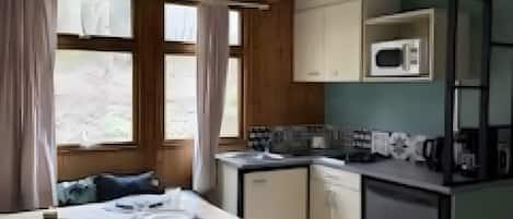 A compact kitchen with a microwave, sink, and cabinets. There is a dining table with chairs and a window with curtains.