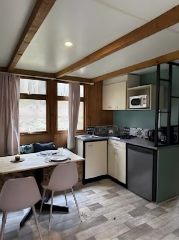 A compact kitchen with a microwave, sink, and cabinets. There is a dining table with chairs and a window with curtains.